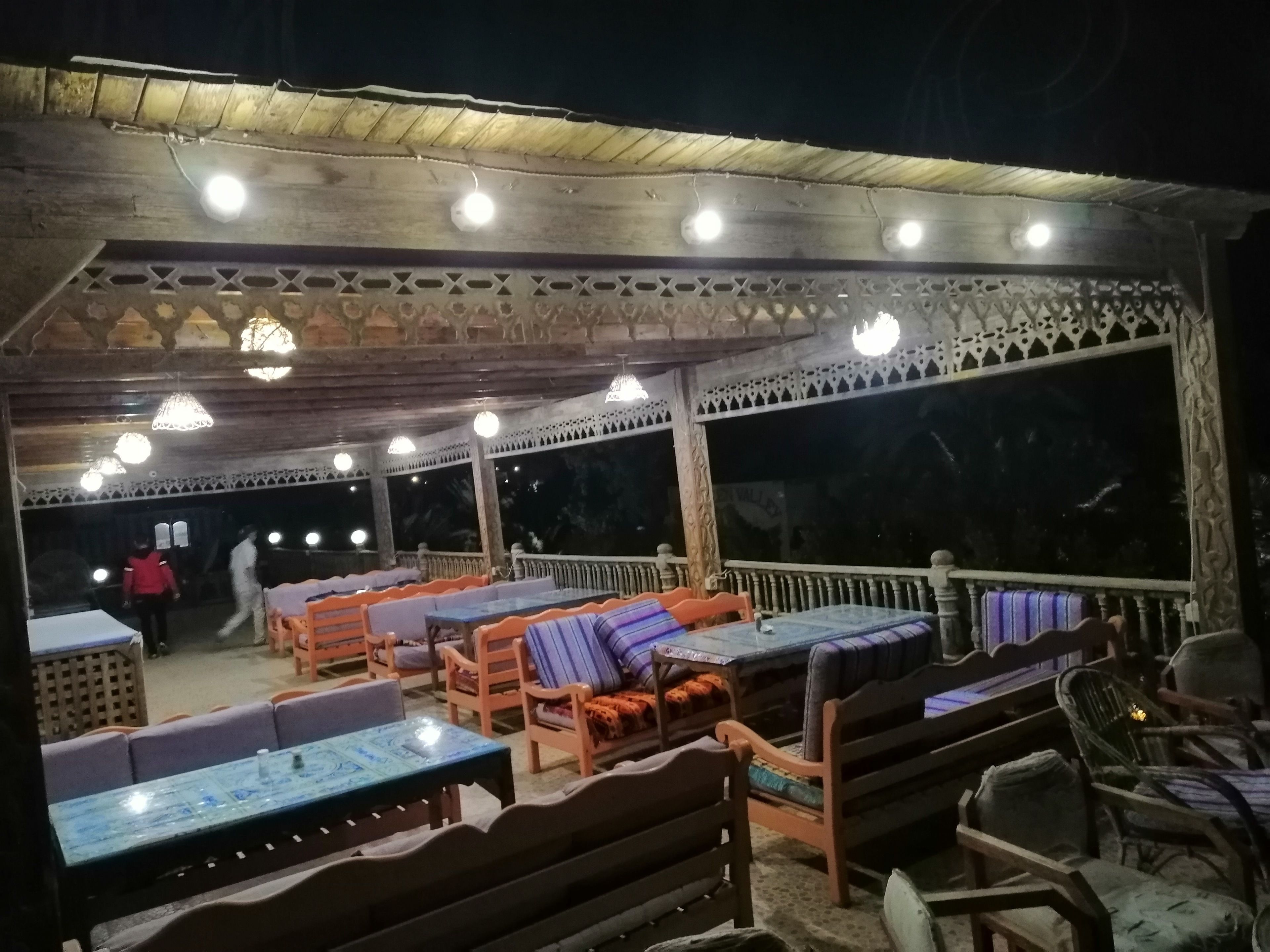 An outdoor seating area with tables and benches, under a covered roof with decorative railings and lighting.