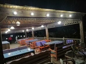 An outdoor seating area with tables and benches, under a covered roof with decorative railings and lighting.