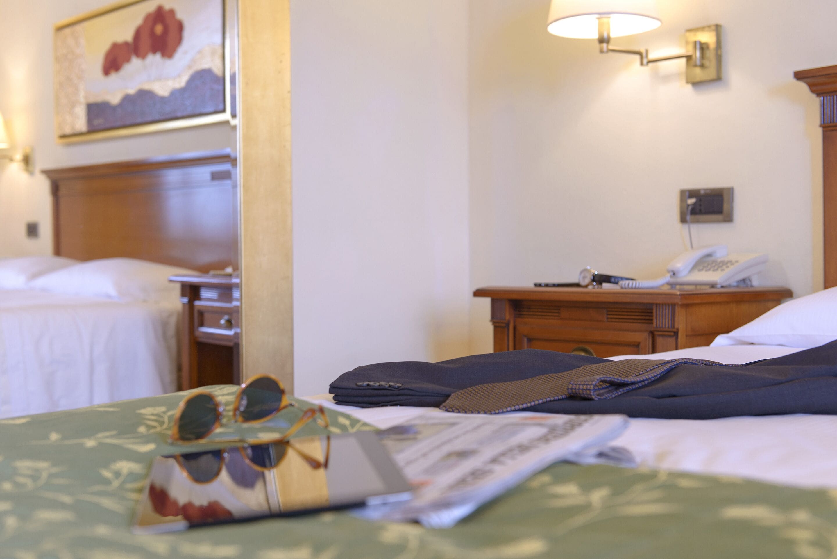 A hotel room with a neatly made bed, a bedside table with a lamp, a telephone, and a painting on the wall.