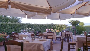 An outdoor dining area with tables and chairs under large umbrellas, surrounded by greenery and flowers. - Popilia Country Resort (Maierato)