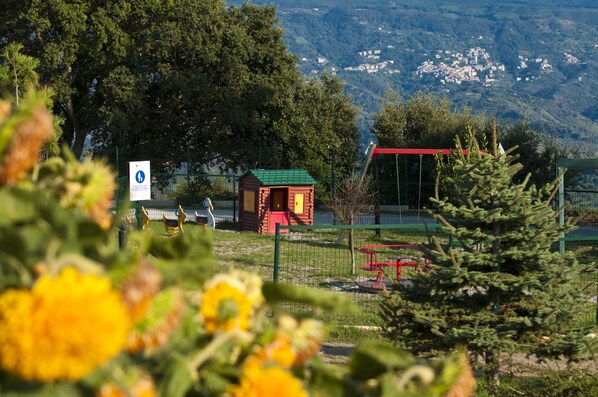 A playground with a red playhouse and a green fence, surrounded by trees and flowers. - Popilia Country Resort (Maierato)