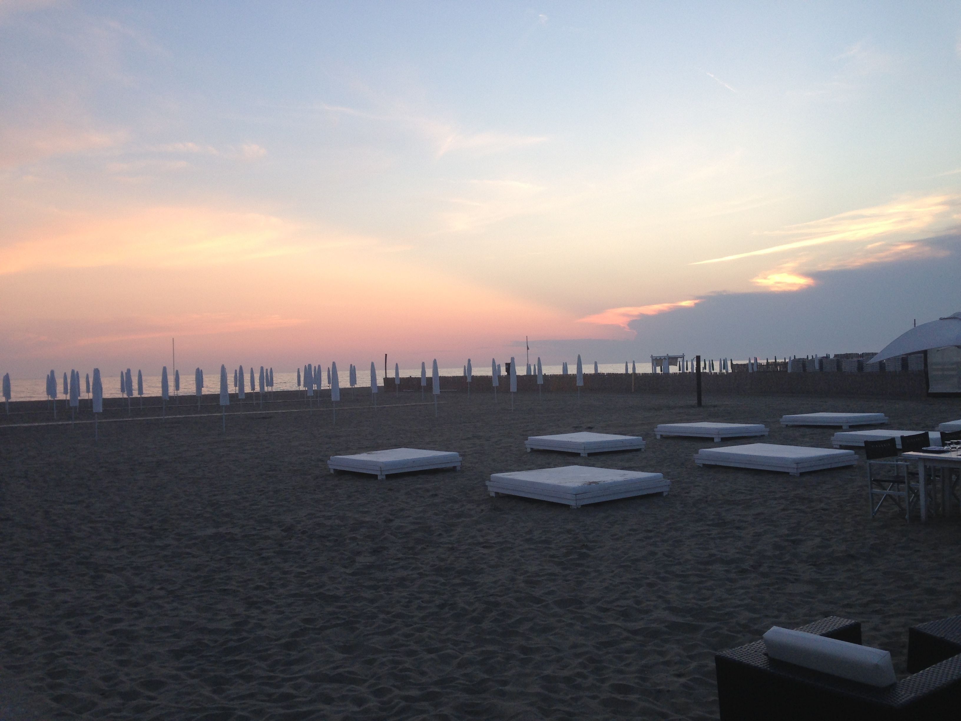 Una playa con hileras de sillas reclinables blancas y sombrillas, algunos colchones en la arena, y una vista despejada del océano.