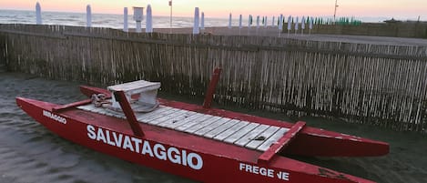 A red boat named 'SALVATAGGIO' on a sandy beach with a wooden fence and flags in the background.