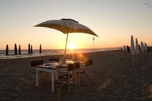 Un coin repas aménagé sur la plage, avec une table et des chaises sous un parasol, offrant une vue sur l’océan au coucher du soleil.