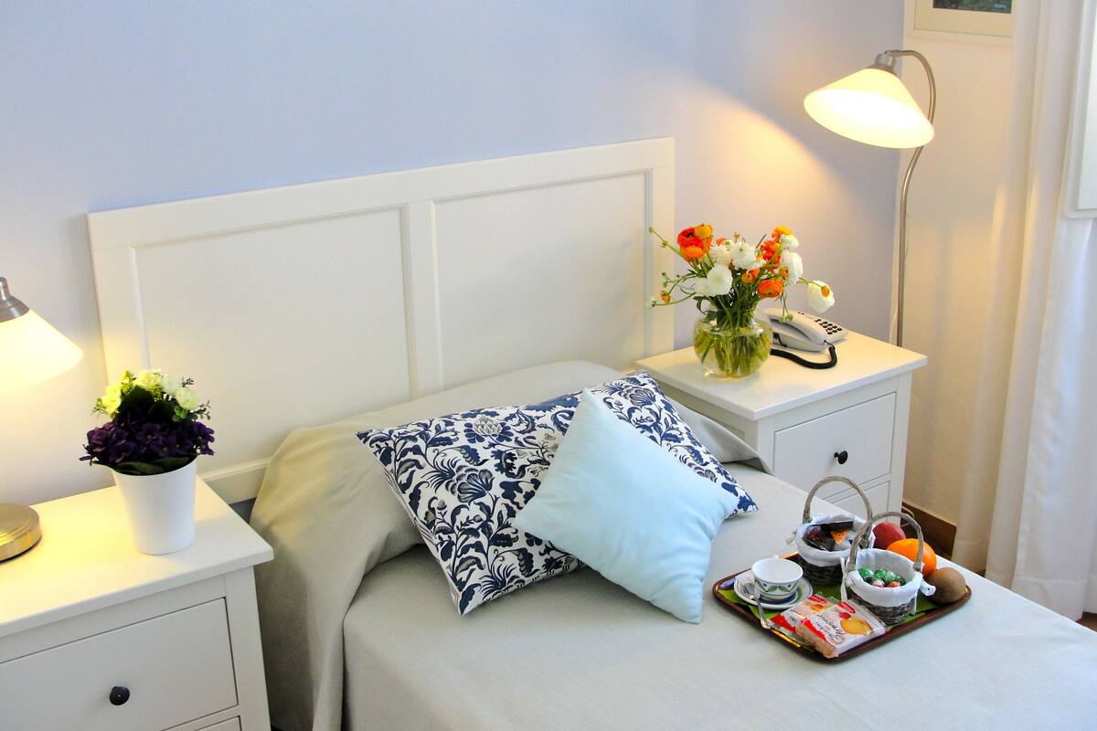 Un lit bien fait, avec une tête de lit blanche, une table de chevet sur laquelle se trouve une lampe, un vase rempli de fleurs et un plateau contenant des éléments pour le petit-déjeuner.