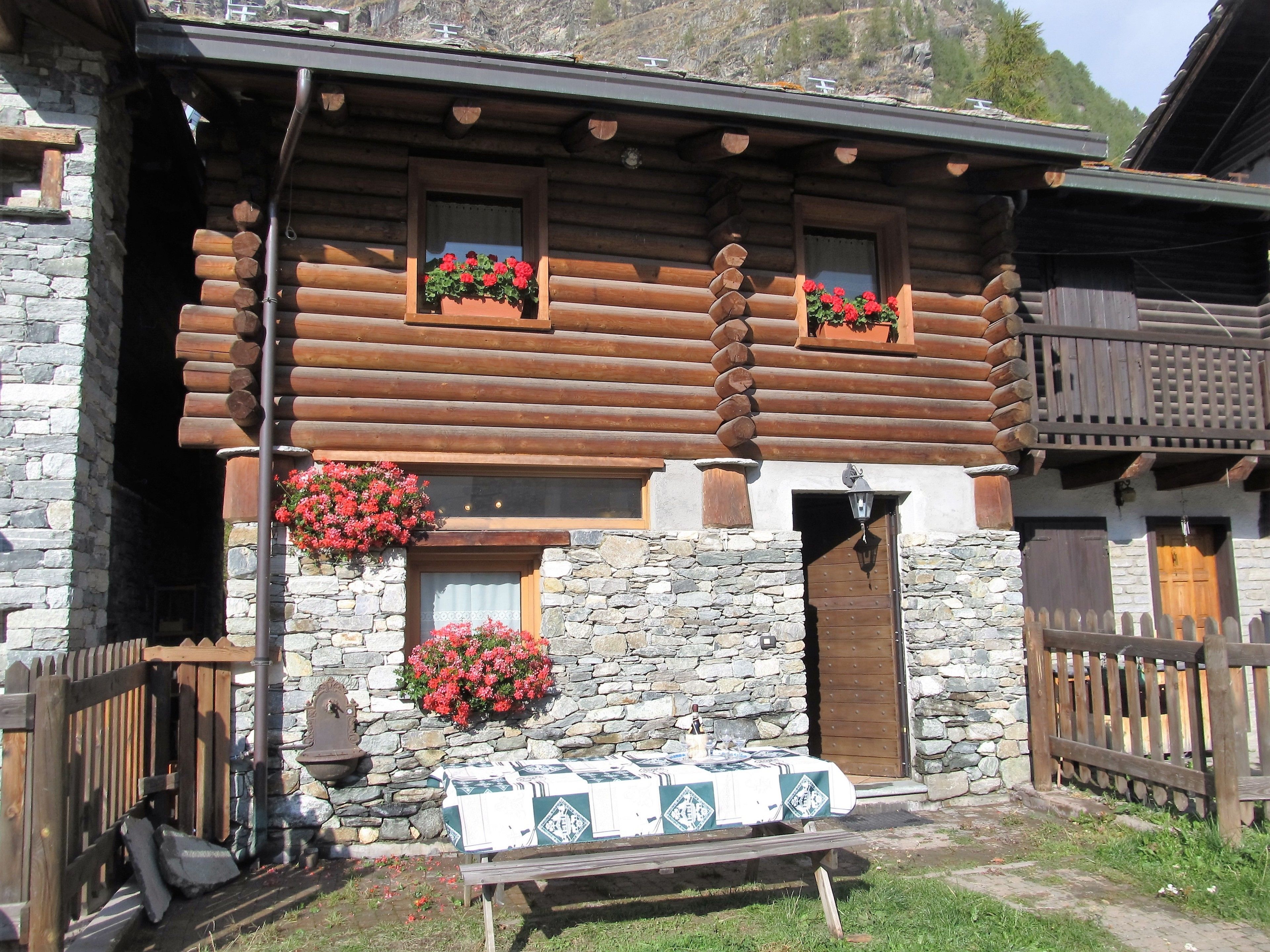 Une cabane en rondins avec des murs en pierre, des fenêtres en bois ornées de jardinières et une table dressée à l’extérieur.
