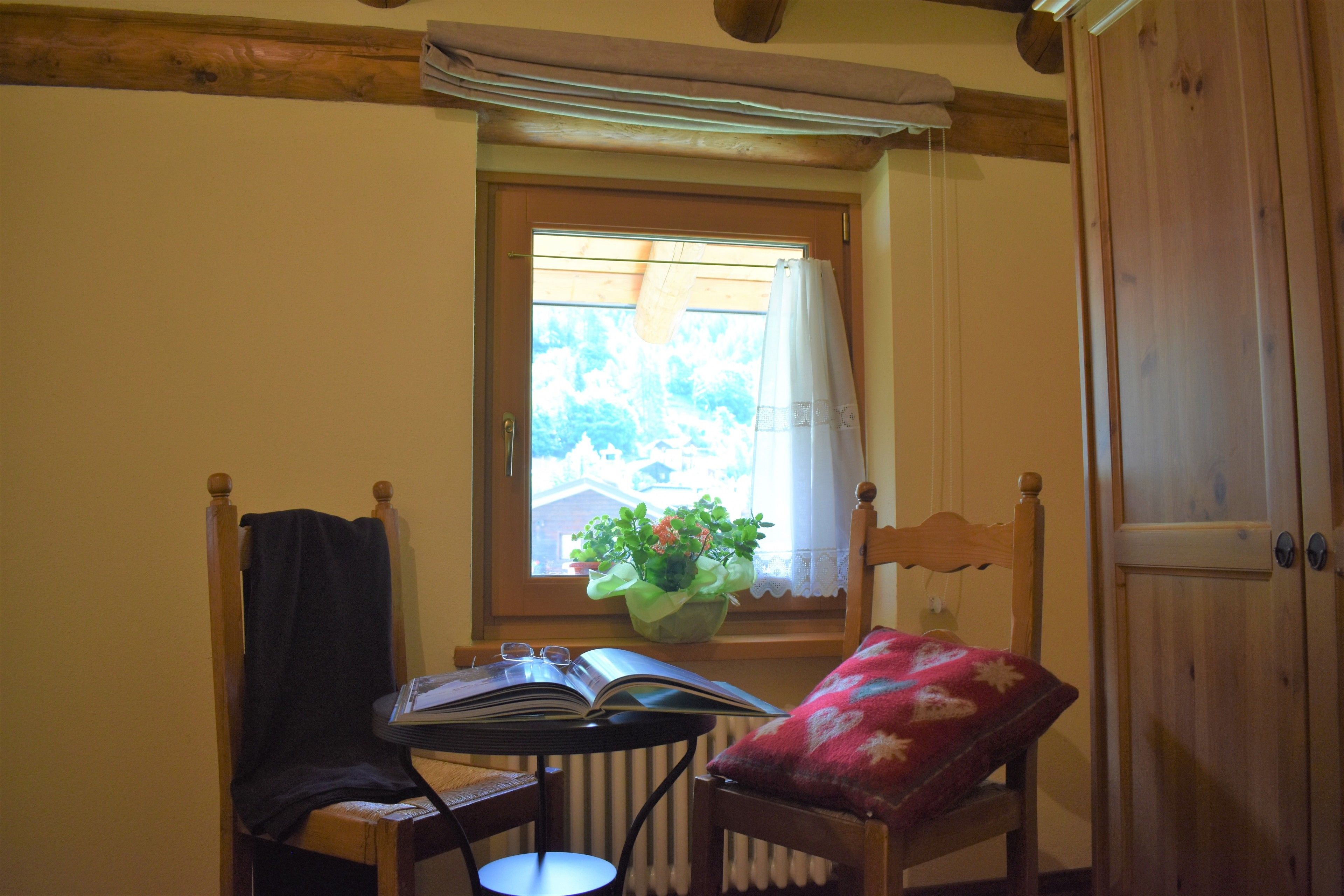 A cozy room with a wooden chair, a small table with an open book, a potted plant, and a window with a view of greenery.