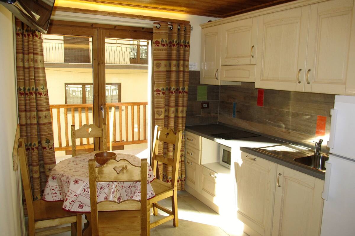 A kitchen with wooden cabinets, a dining table with chairs, and a balcony with a wooden railing.