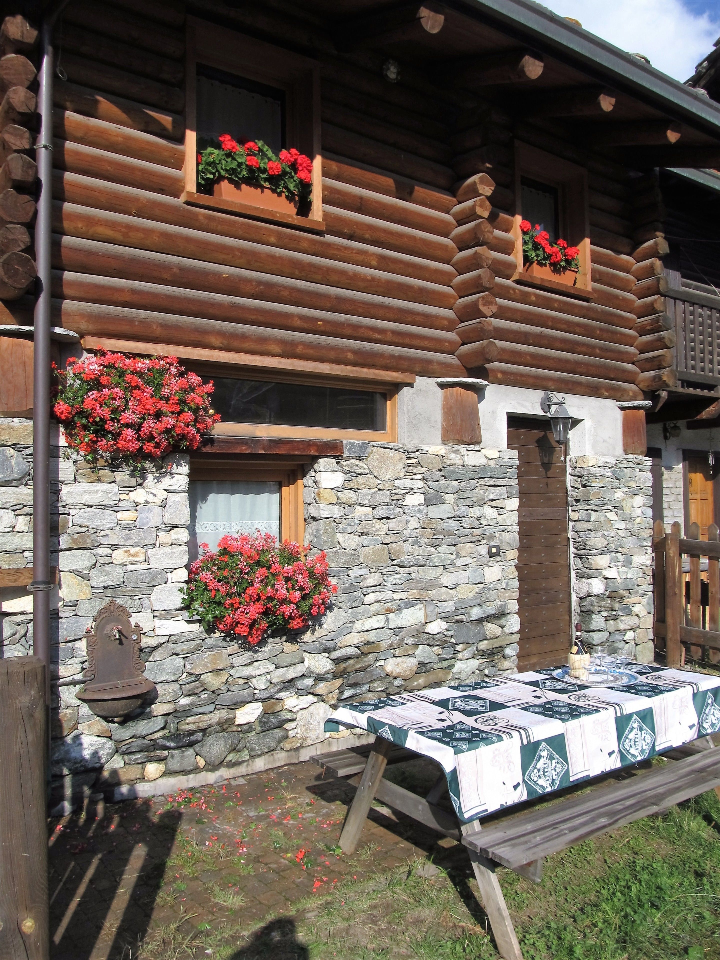 A log cabin with stone walls, flower boxes with red flowers, and a table set for dining.
