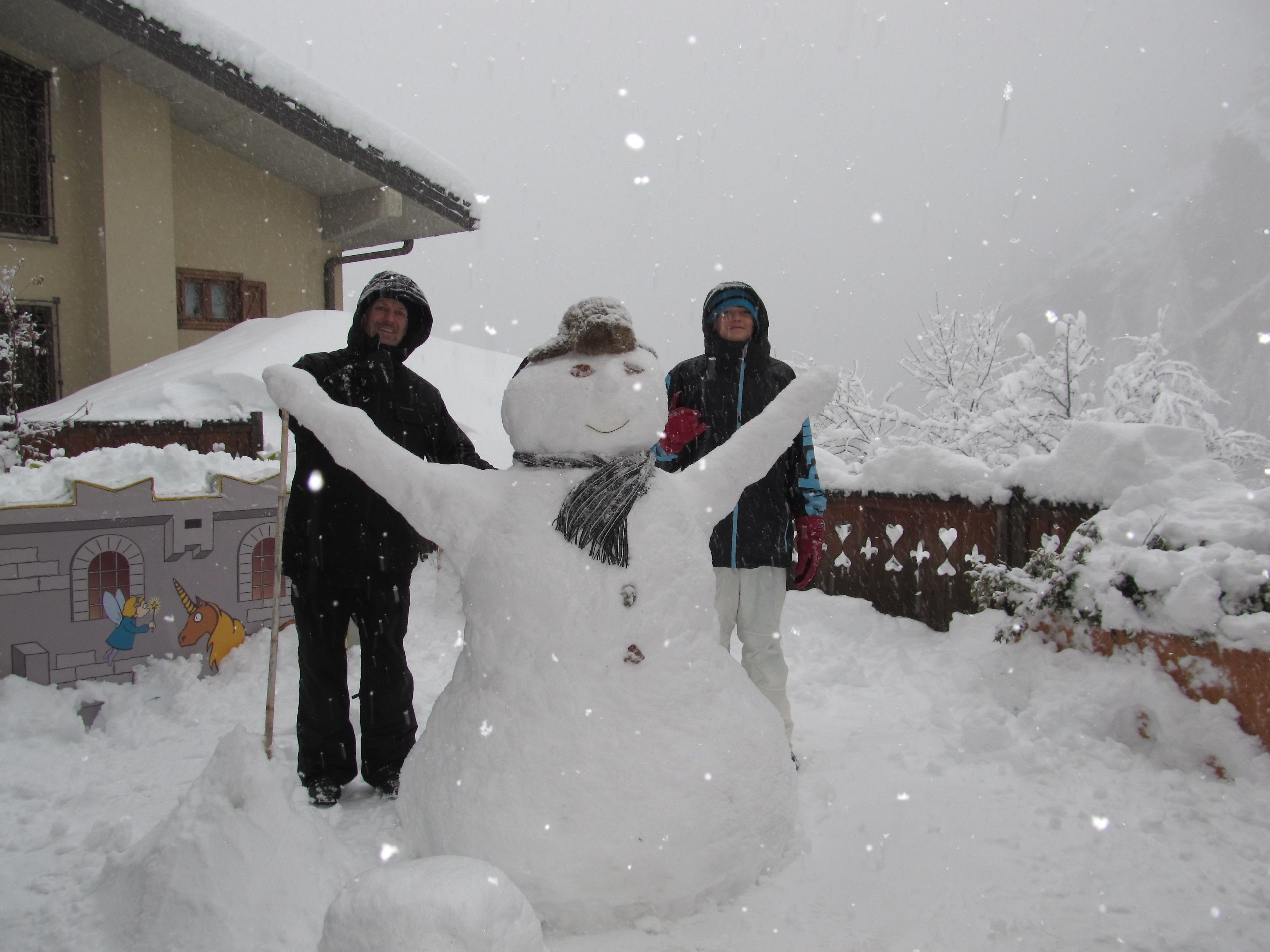 Ein Schneemann, neben dem zwei Personen stehen, ein Haus mit schneebedecktem Dach und eine verschneite Landschaft.