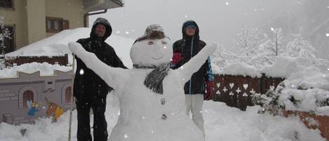 Ein Schneemann, neben dem zwei Personen stehen, ein Haus mit schneebedecktem Dach und eine verschneite Landschaft.