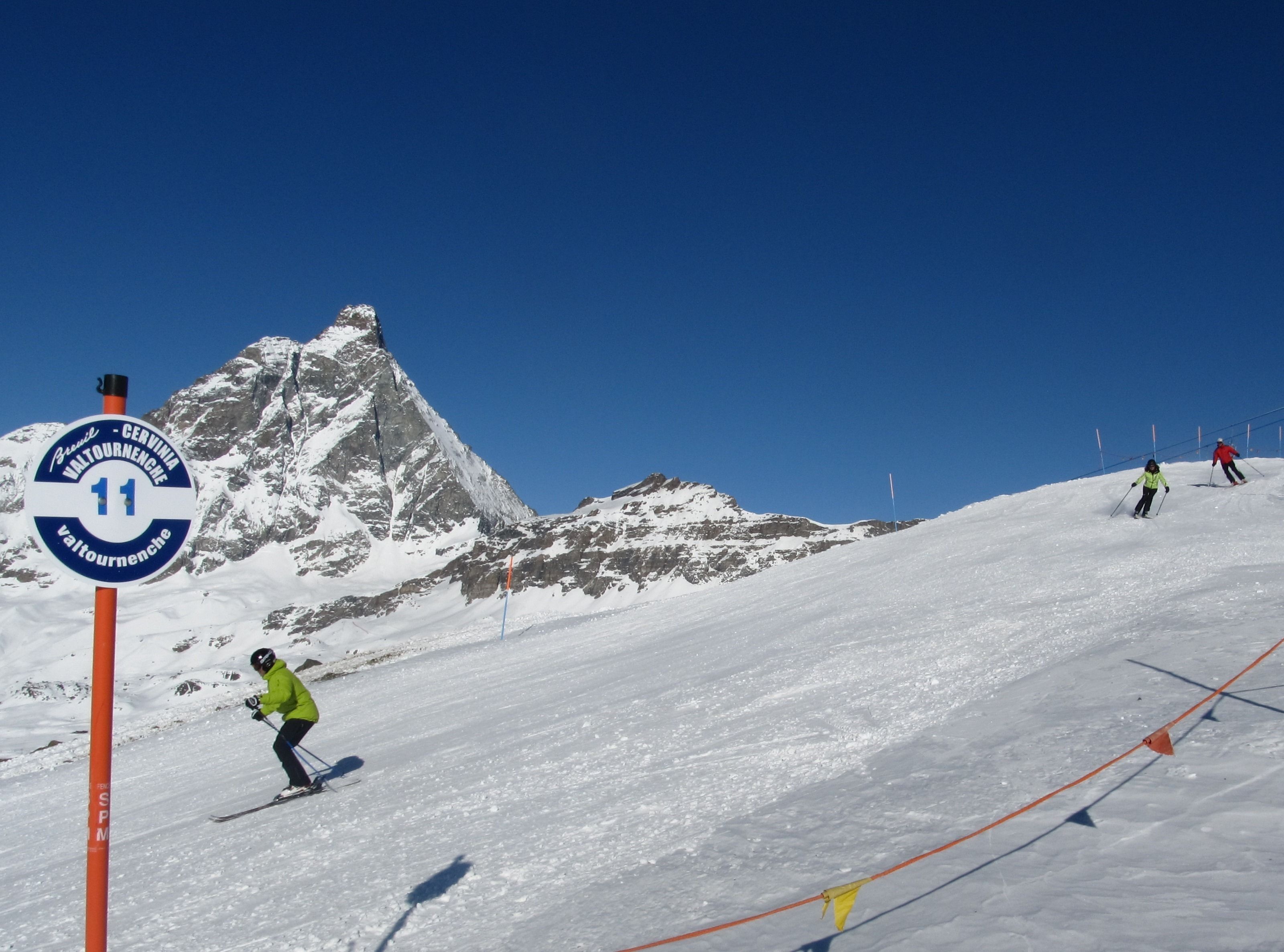 A ski slope with a sign indicating route number 11, skiers in motion, and a mountain backdrop.