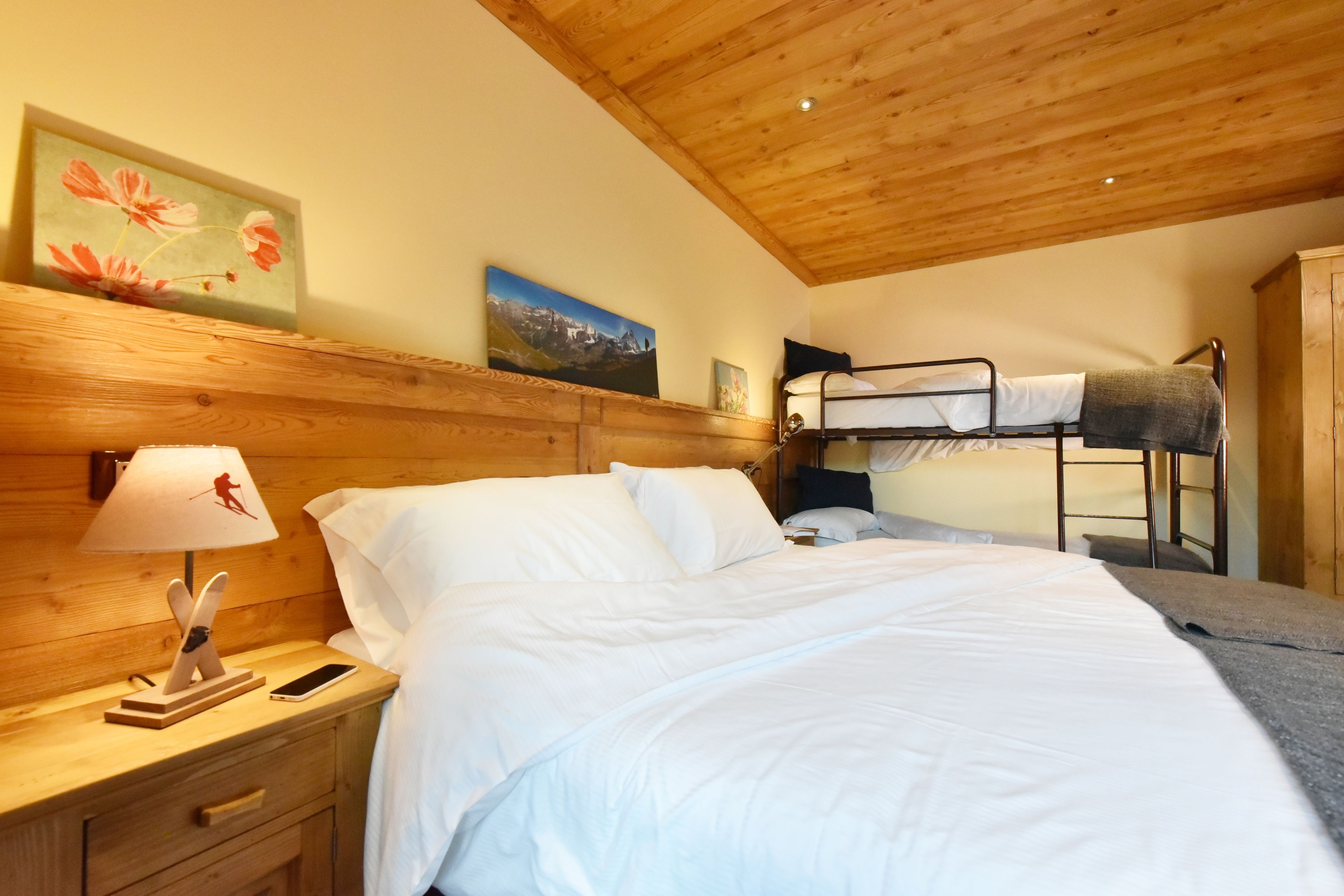 Una habitación acogedora con techo de madera, una litera, una cama individual, una mesita de noche con lámpara y un cuadro en la pared.