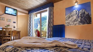 A bedroom with a bed, a television, a painting of mountains, and a view of a mountainous landscape through the window. - Miramonti (Valtournenche)
