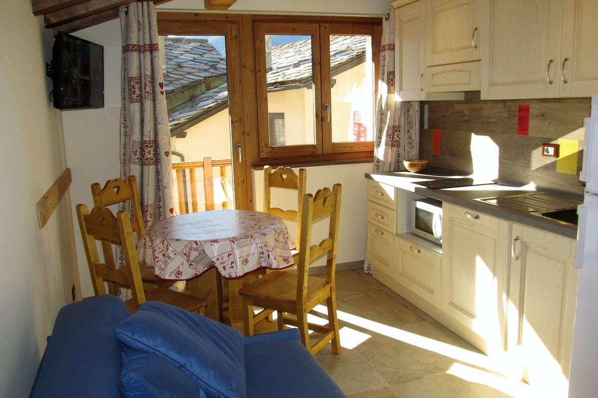 A small kitchen with wooden cabinets, a dining table with chairs, and a TV mounted on the wall.