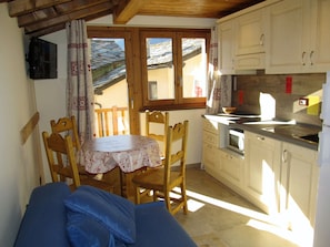 A small kitchen with wooden cabinets, a dining table with chairs, and a TV mounted on the wall. - Miramonti (Valtournenche)