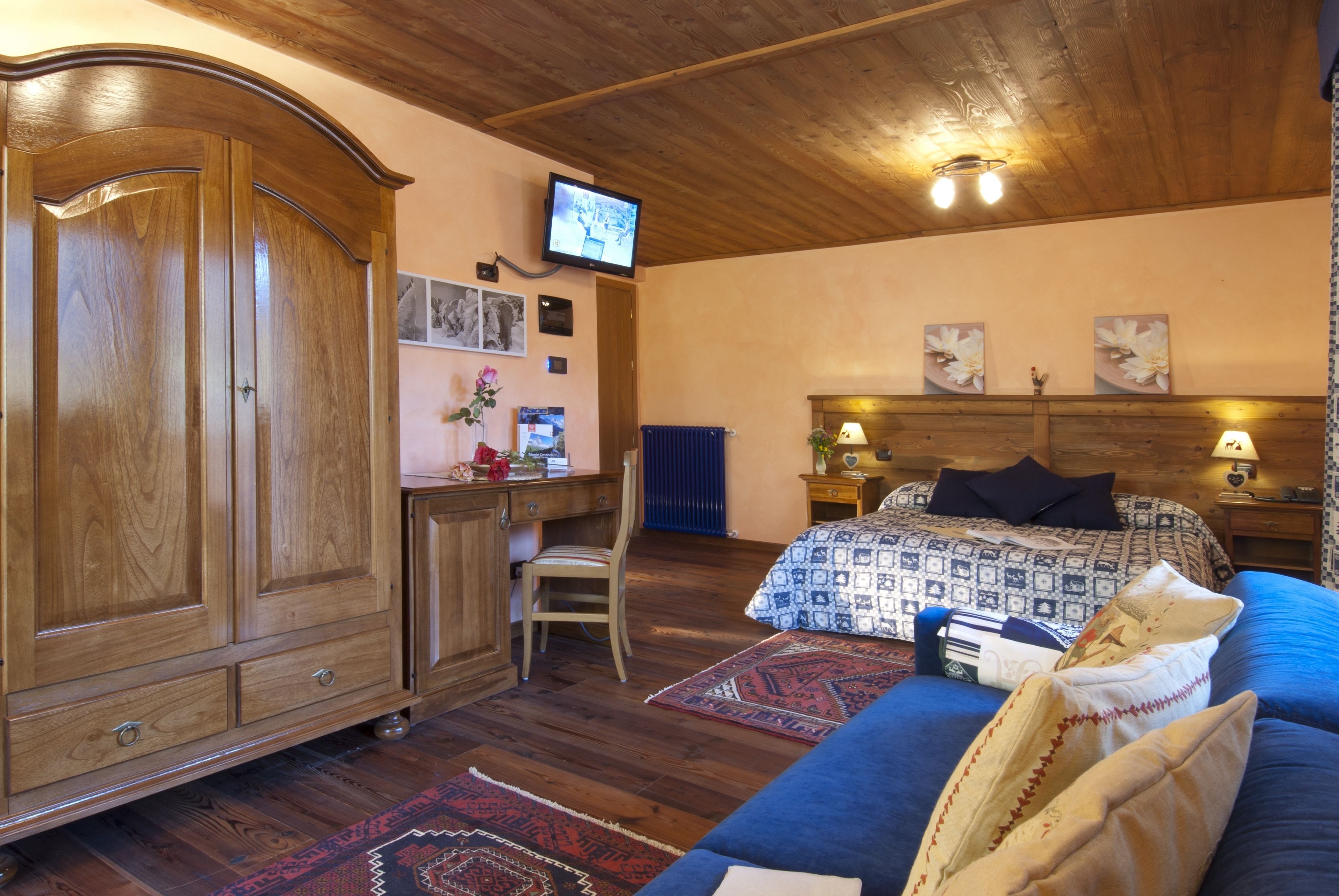 A bedroom with a wooden wardrobe, a desk, a chair, a bed with patterned bedding, and a television on the wall.