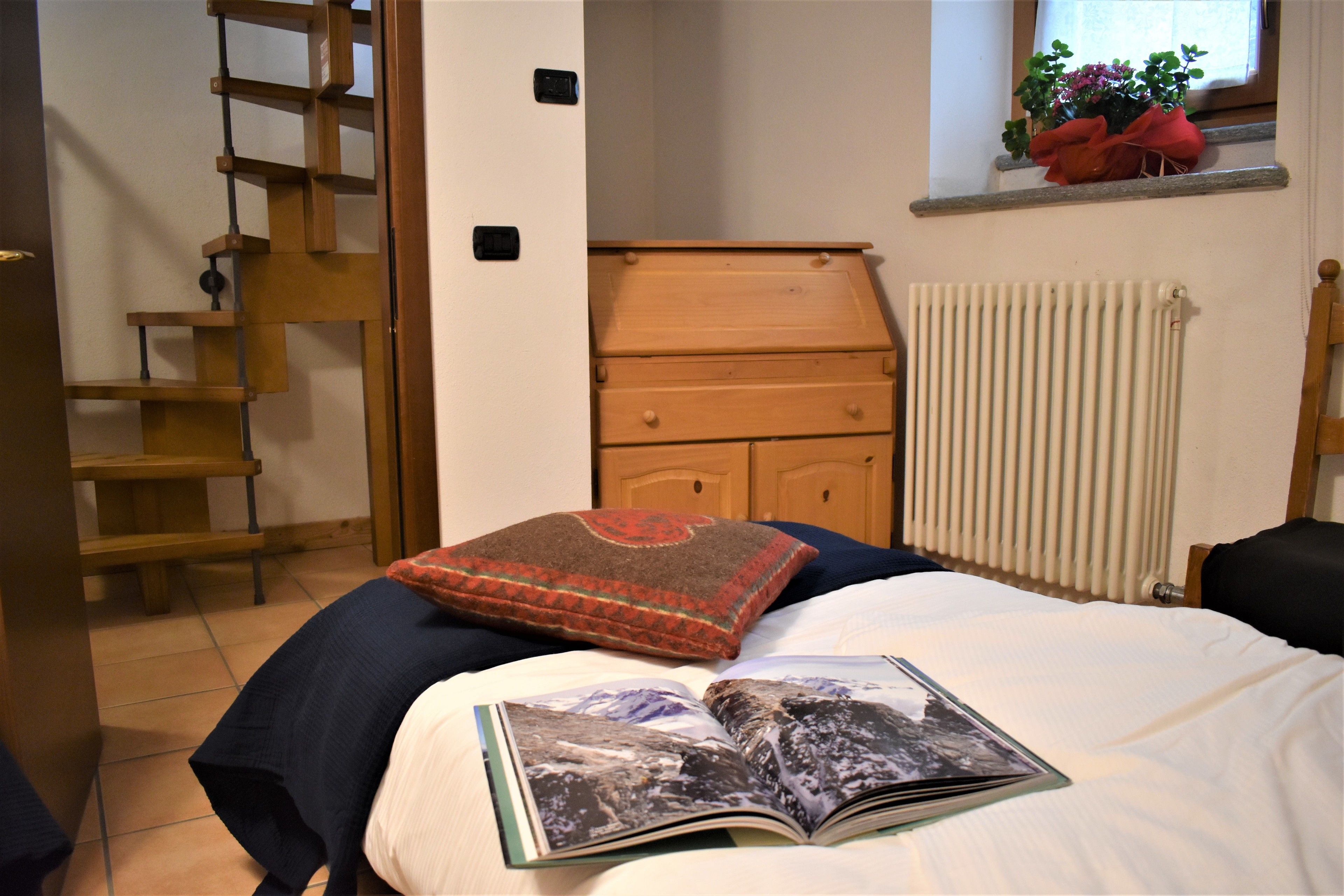 A bedroom with a bed, a wooden ladder, a dresser, a radiator, and a window with a plant.