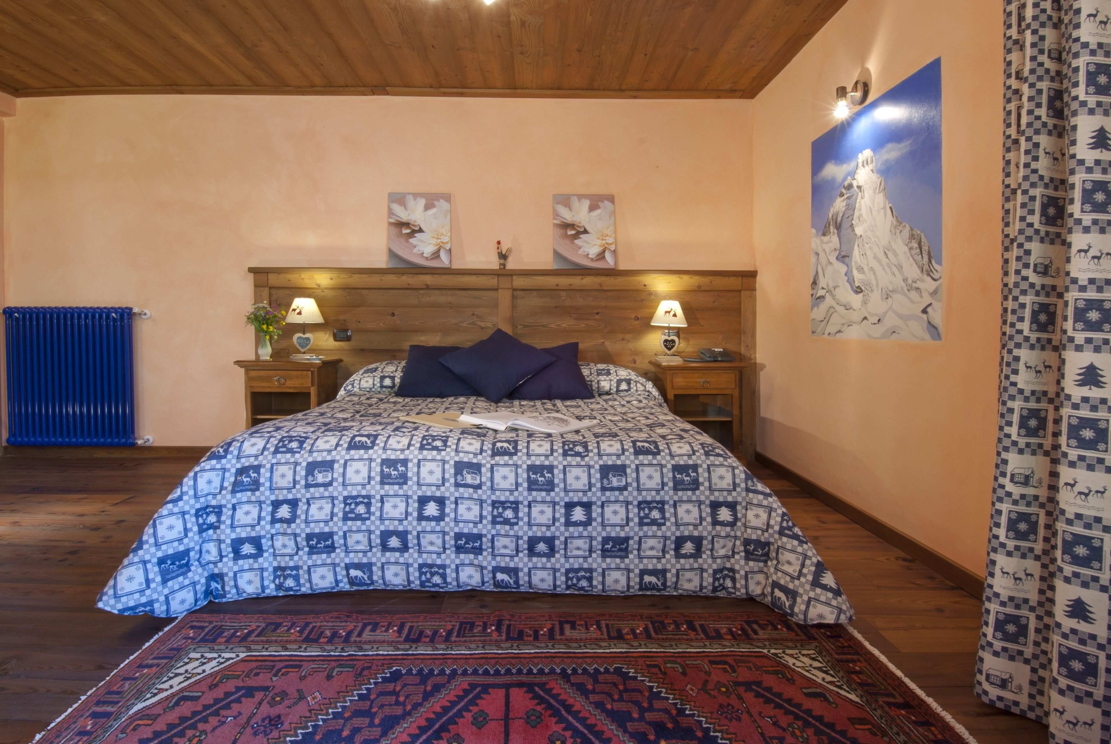 Un dormitorio con edredón estampado, cabecero de madera, dos lámparas fijadas a la pared, un radiador azul y un cuadro grande con un paisaje de montaña.