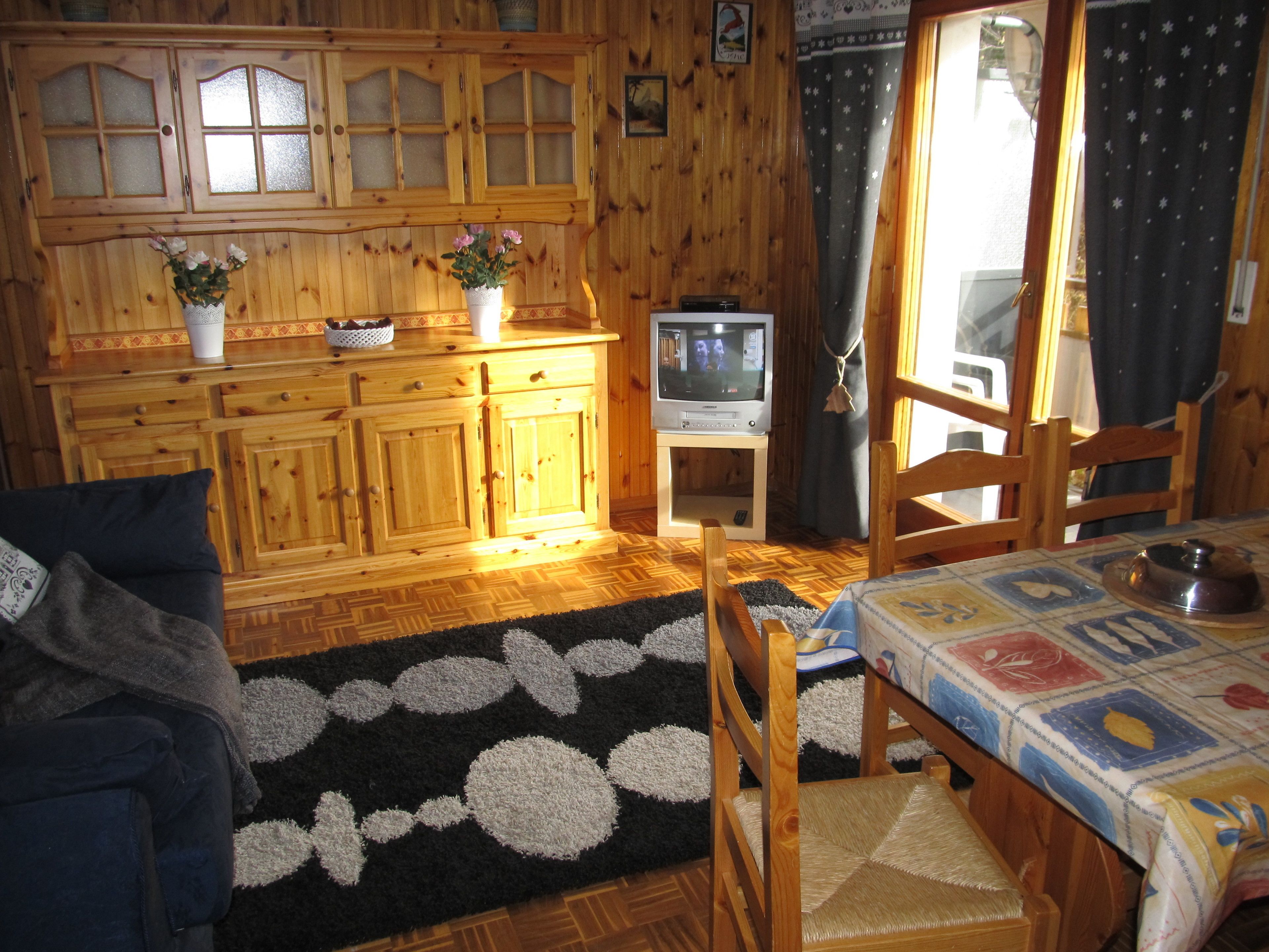 A cozy wooden cabin interior with a sofa, a table with a patterned tablecloth, a television on a stand, and a window with curtains.