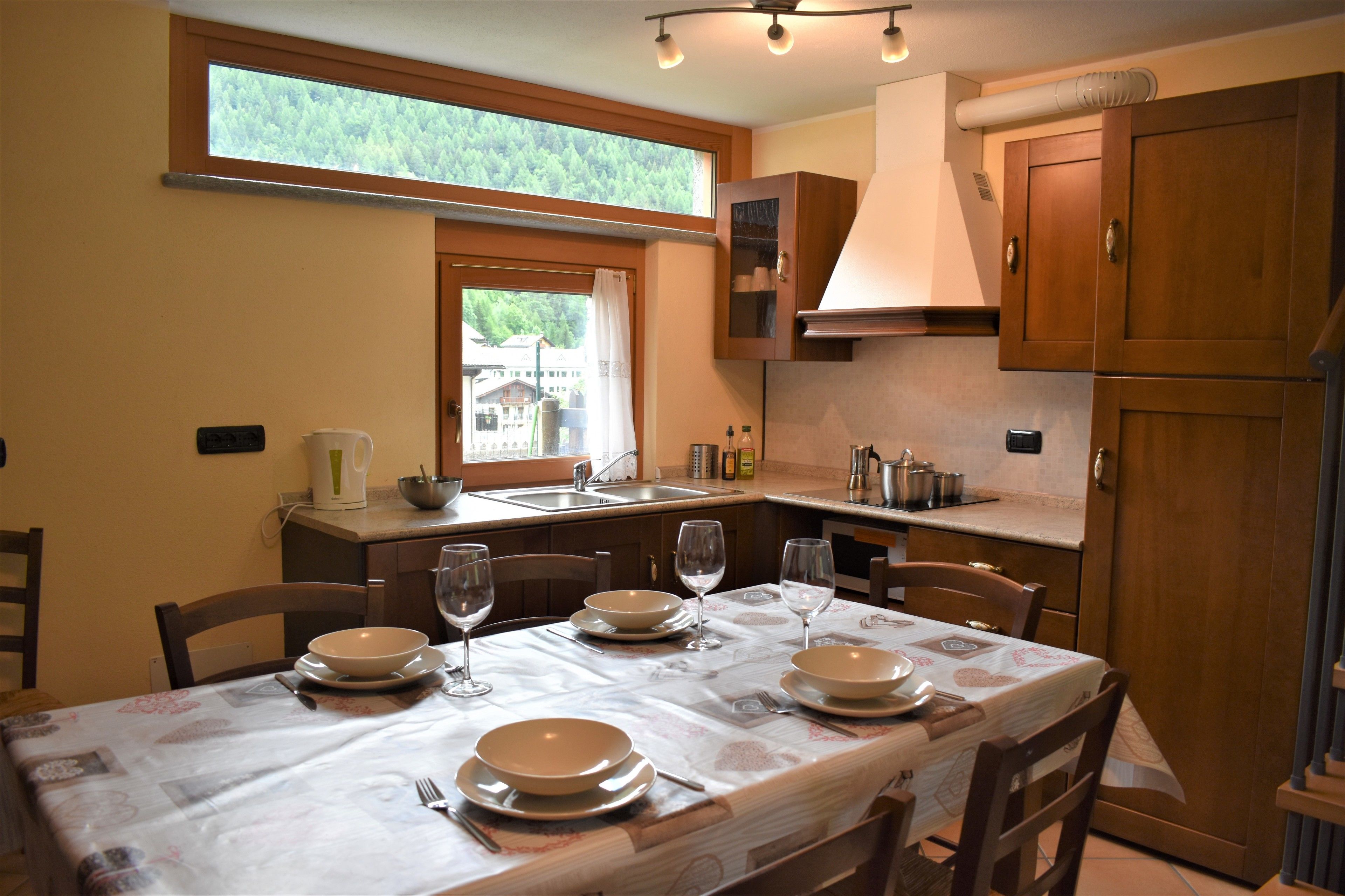Un coin repas avec une table dressée pour quatre personnes, une fenêtre donnant sur un paysage verdoyant, des meubles de rangement en bois et un espace cuisine équipé d’un évier et d’une cuisinière.