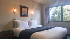 A hotel room with a bed, a nightstand, a painting, a window with curtains, and a view of trees outside. - Aspen Lodge Motel (Hanmer Springs)