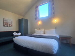 Habitación de hotel con cama, mesita de noche, armario, un cuadro en la pared y ventana con cortinas.