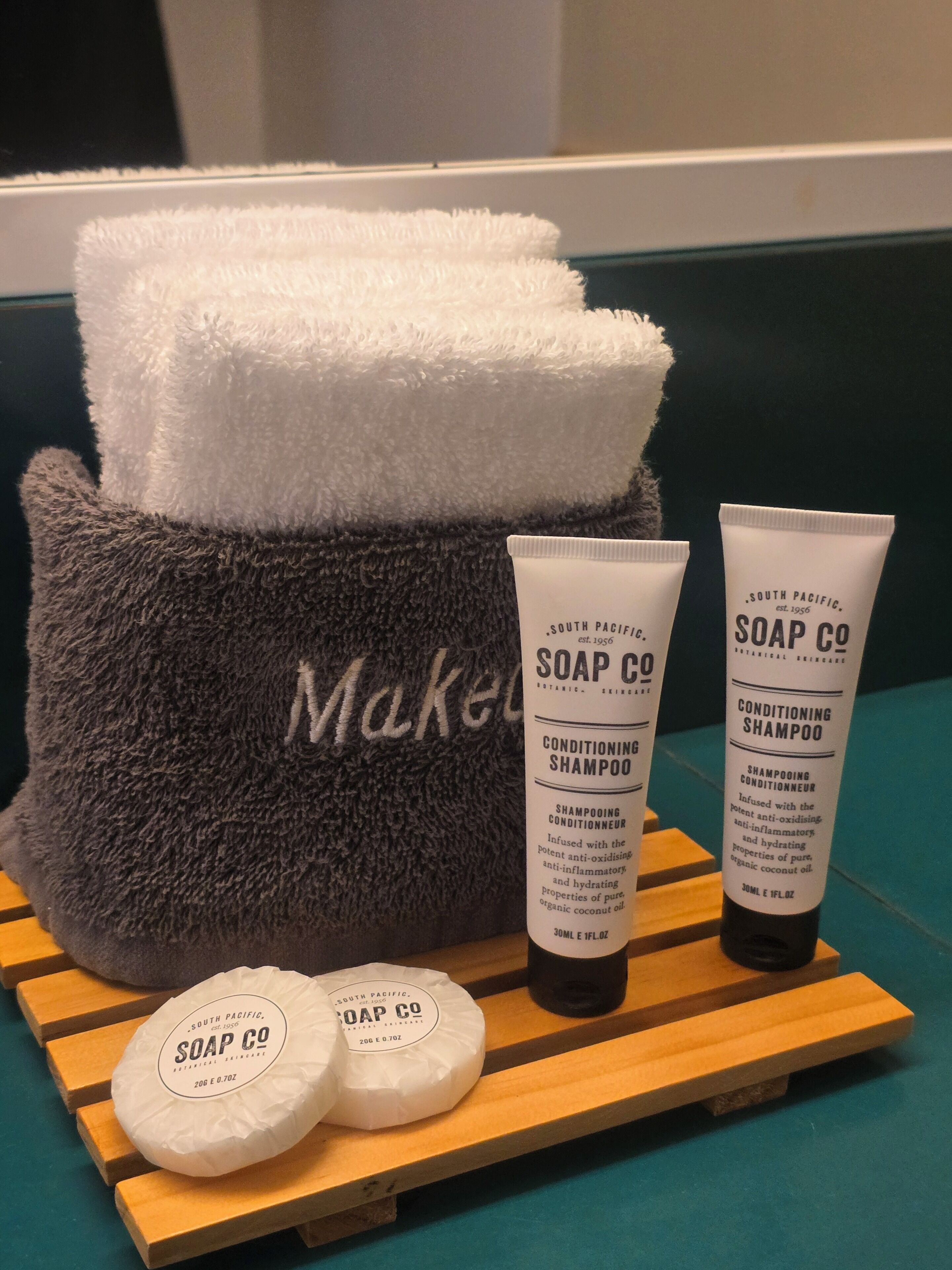 A grey towel with 'Make' embroidered, two soap bottles, and a bar of soap on a wooden surface.