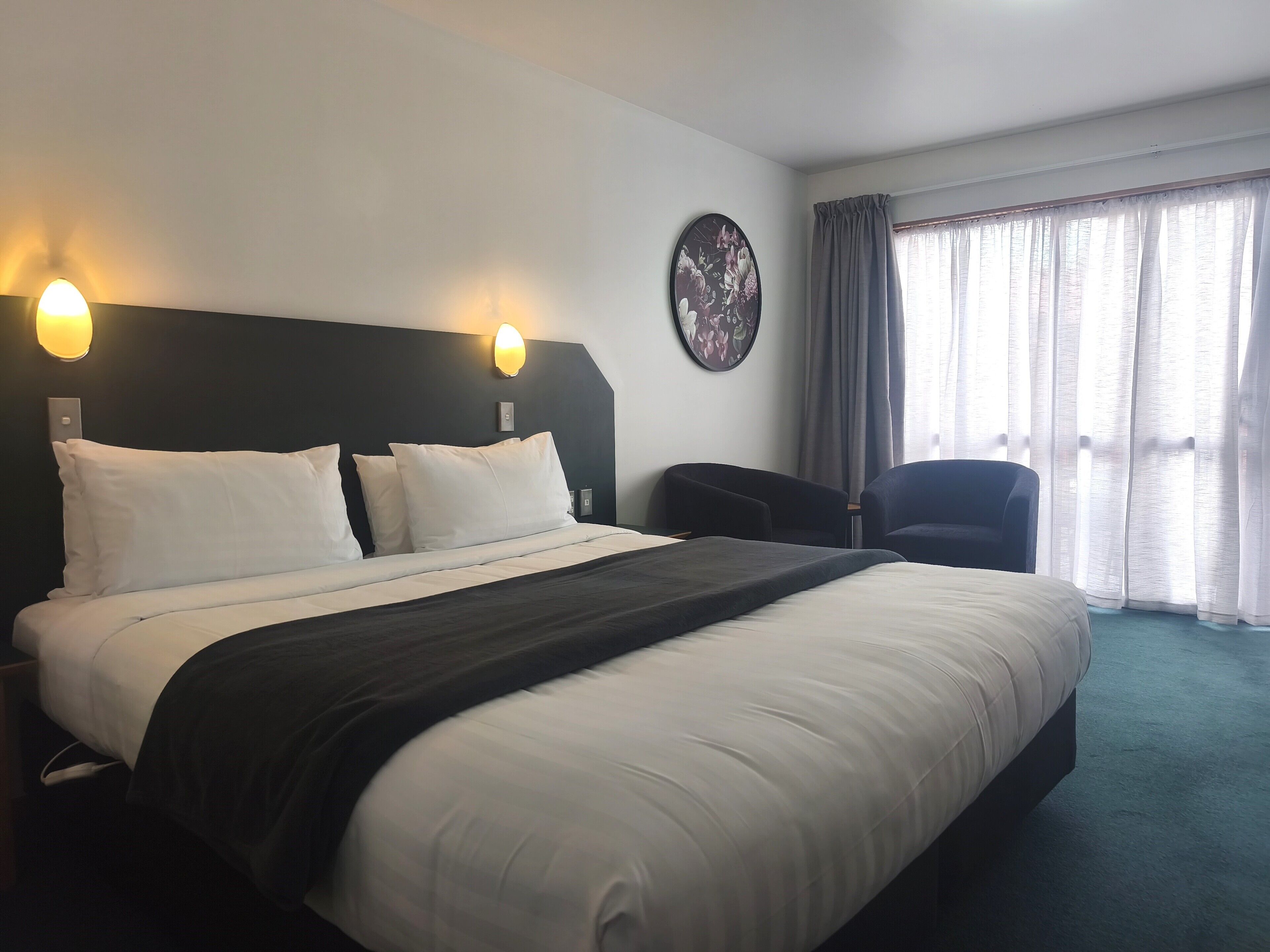 Habitación de hotel con una cama grande, dos lámparas de pared, un adorno circular en la pared, una silla y una ventana con cortinas.