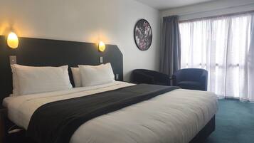 Habitació d'hotel amb un llit gran, dues làmpades penjades a la paret, un objecte decoratiu rodó, una cadira i una finestra amb cortines.