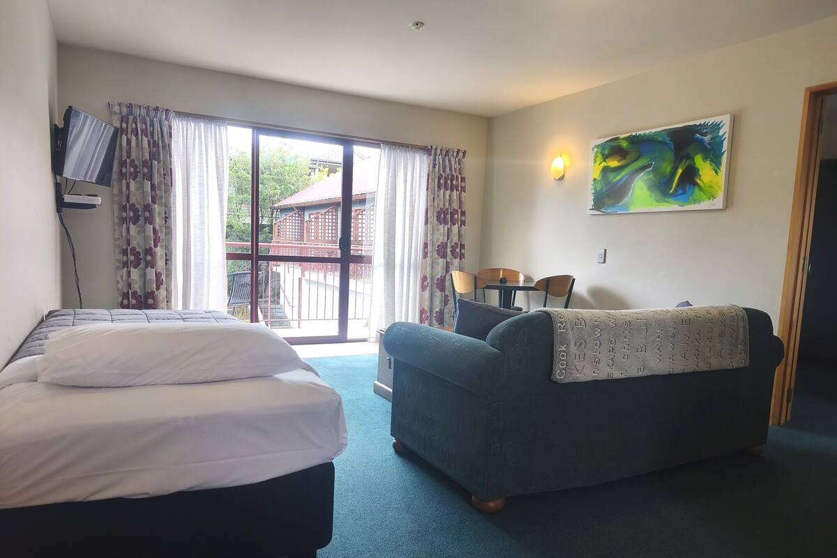 Habitación de hotel con cama, sofá, mesa de comedor con sillas, un cuadro en la pared y vista a un edificio.