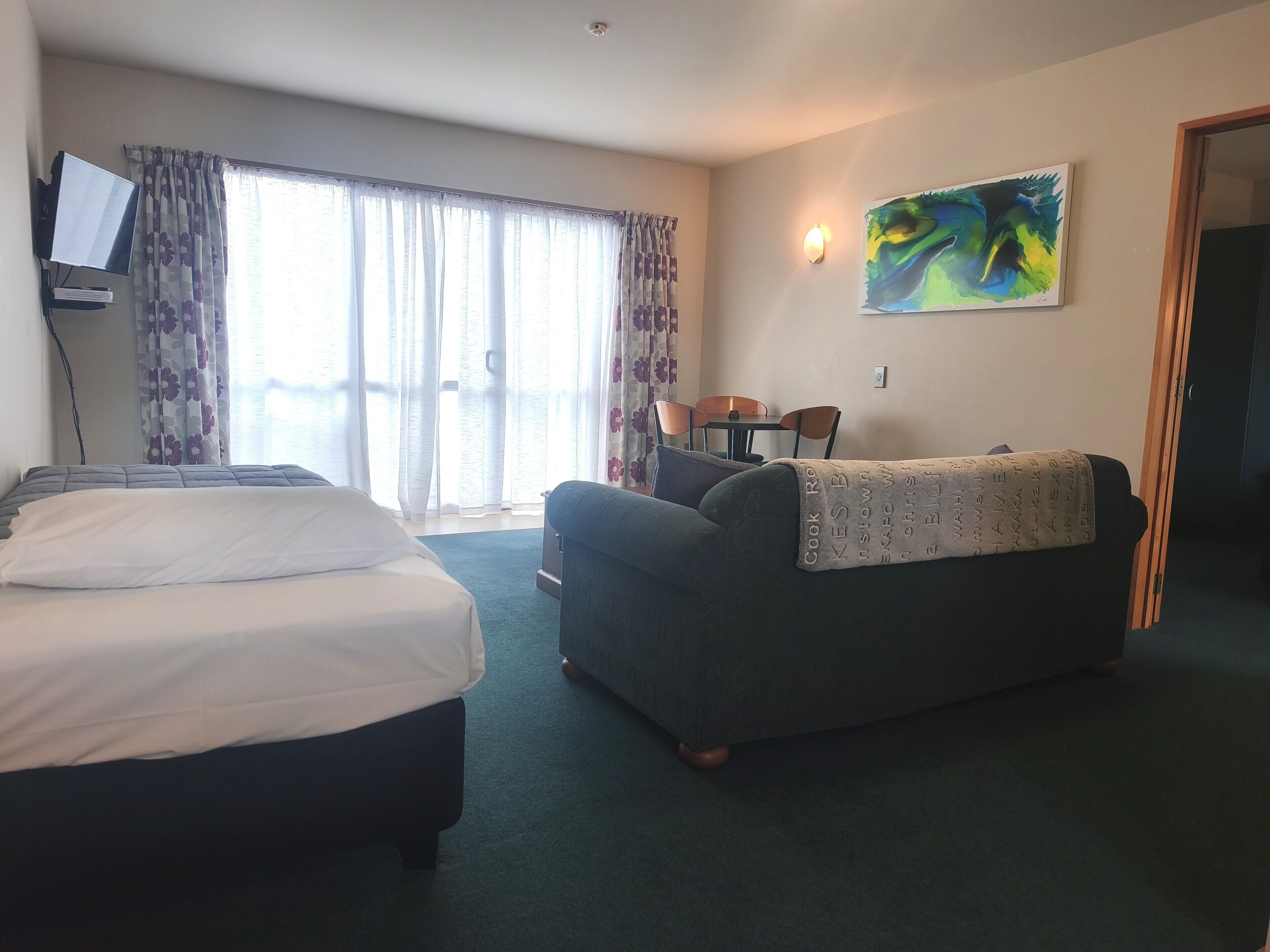 A hotel room with a bed, a sofa, a TV, a painting, and a window with curtains.