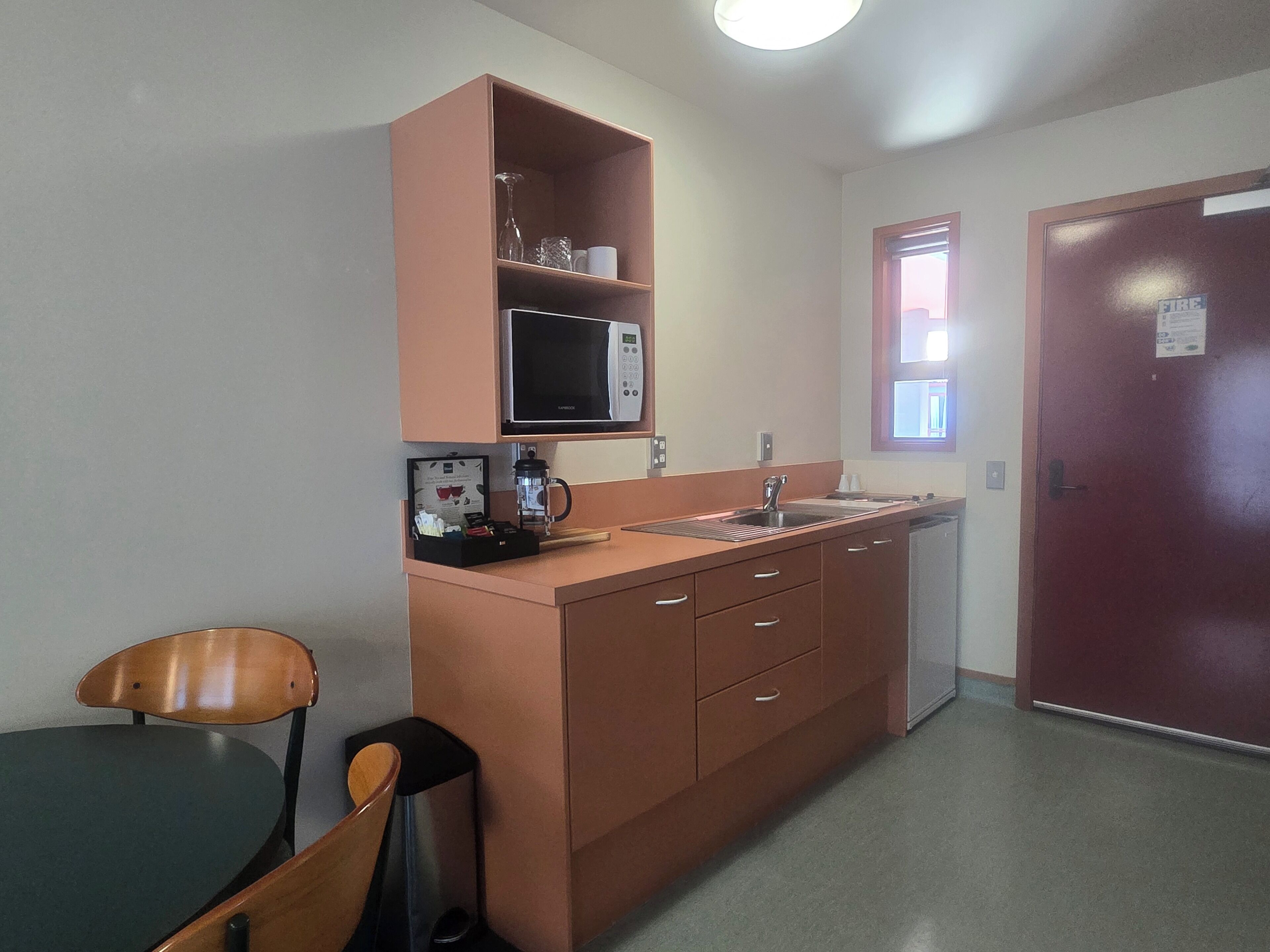 A compact kitchenette with a microwave, coffee maker, and sink. There is a chair and a small table, and a door with a fire safety sign.