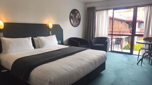 A hotel room with a bed, two chairs, a table, a clock on the wall, and a view of buildings through the window. - Aspen Lodge Motel (Hanmer Springs)