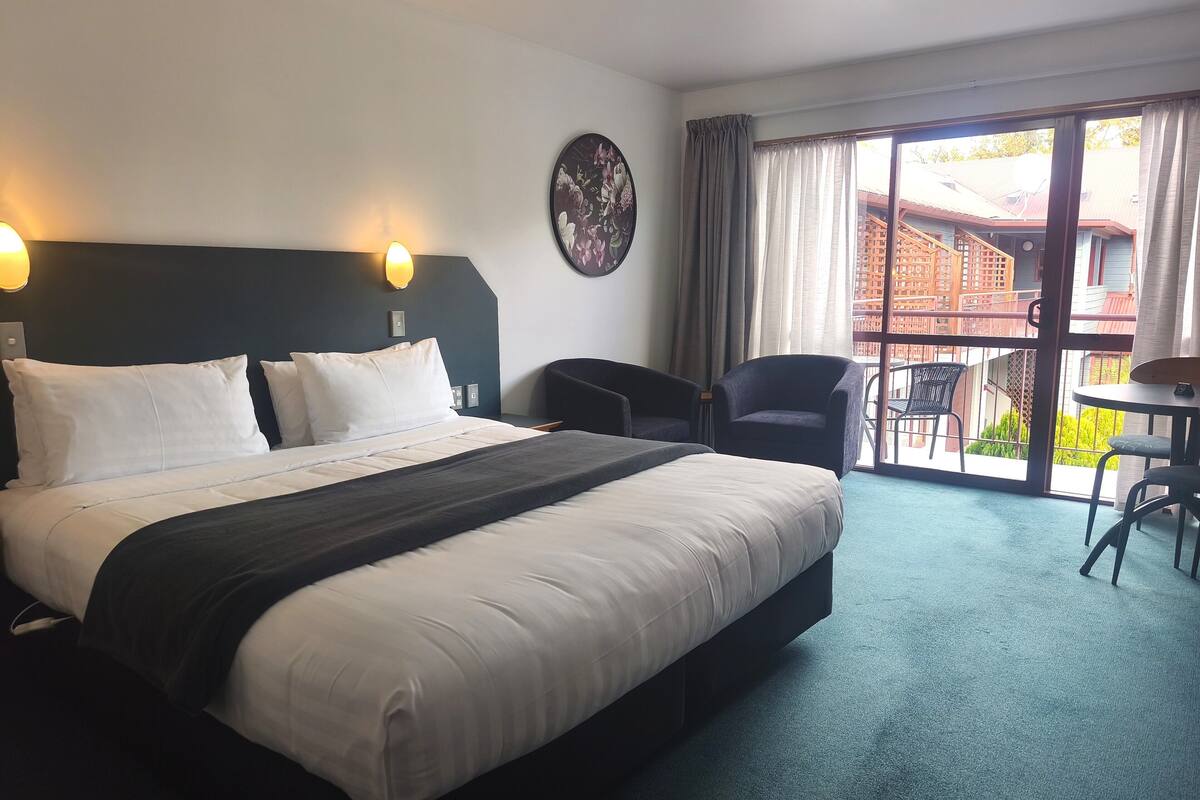 Habitación de hotel con cama, dos sillas, una mesa, un reloj en la pared y vistas a edificios a través de la ventana.