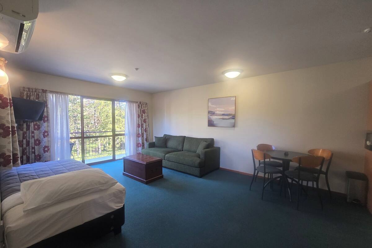 Una habitación con cama, sofá, mesa de comedor con sillas, televisor y ventana con cortinas.