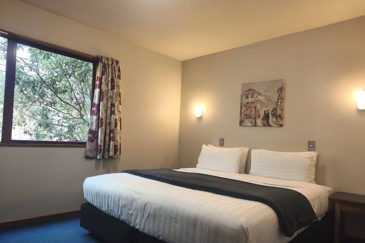 Una habitación de hotel con cama, ventana con cortinas, un cuadro en la pared y una mesita de noche.