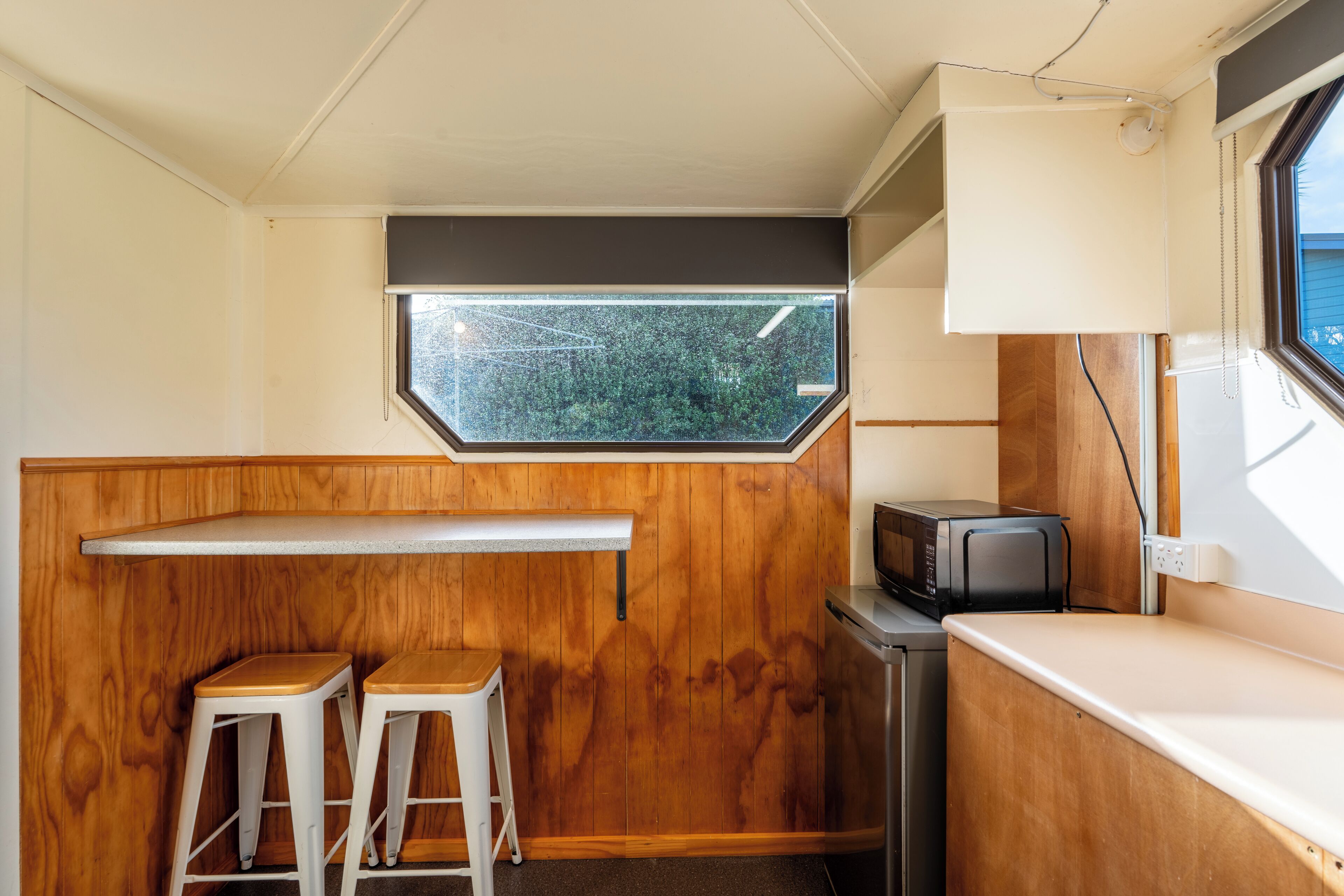 Una kitchenette compacta con encimera de madera, dos taburetes, un microondas y una ventana con vistas a la vegetación.