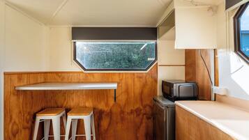 A compact kitchenette with a wooden countertop, two stools, a microwave, and a window providing a view of greenery.