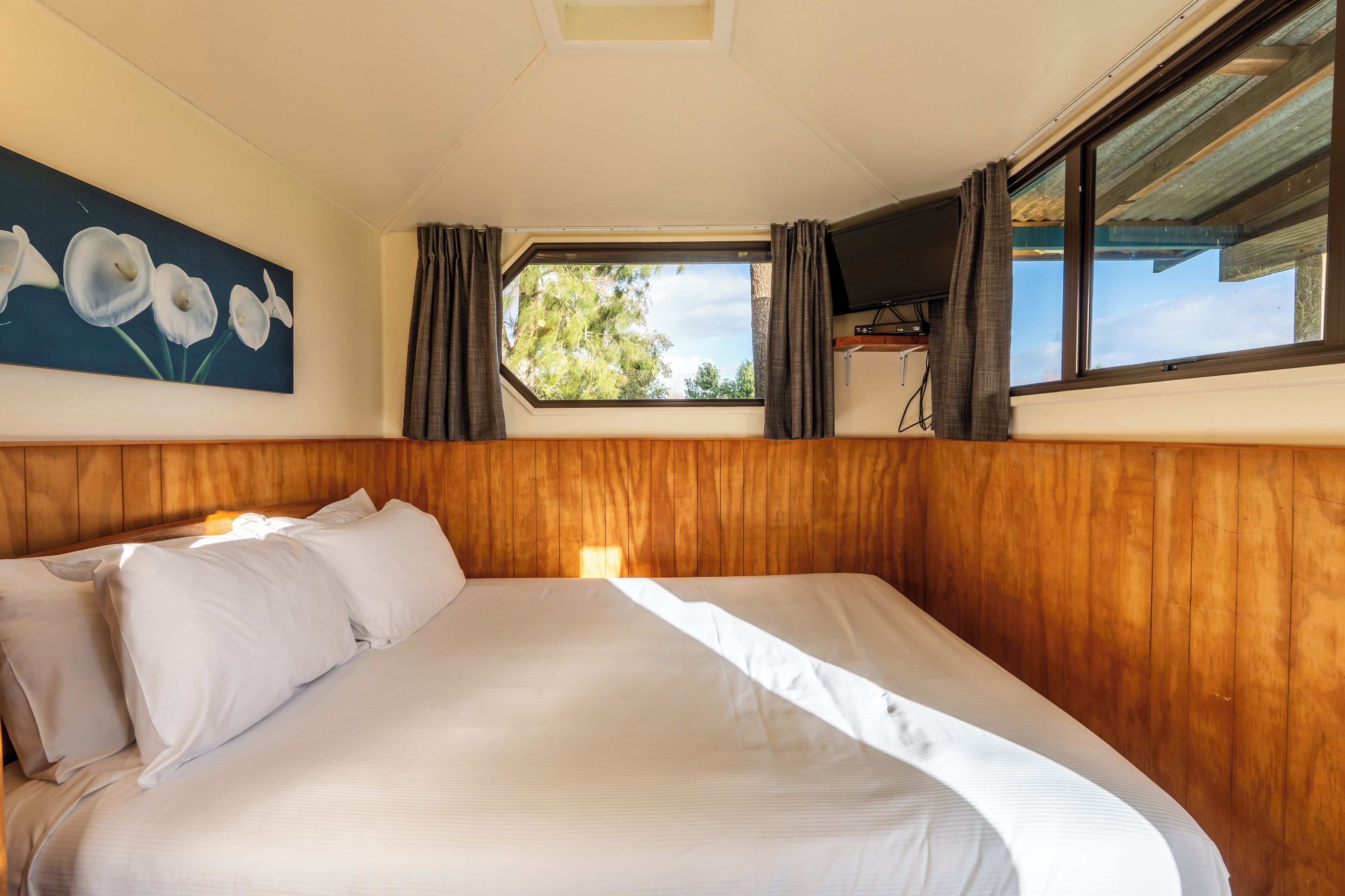 Une chambre à coucher avec une tête de lit en bois, un grand lit recouvert de draps blancs, un tableau représentant des fleurs accroché au mur, une fenêtre avec des rideaux et un téléviseur posé sur un meuble.