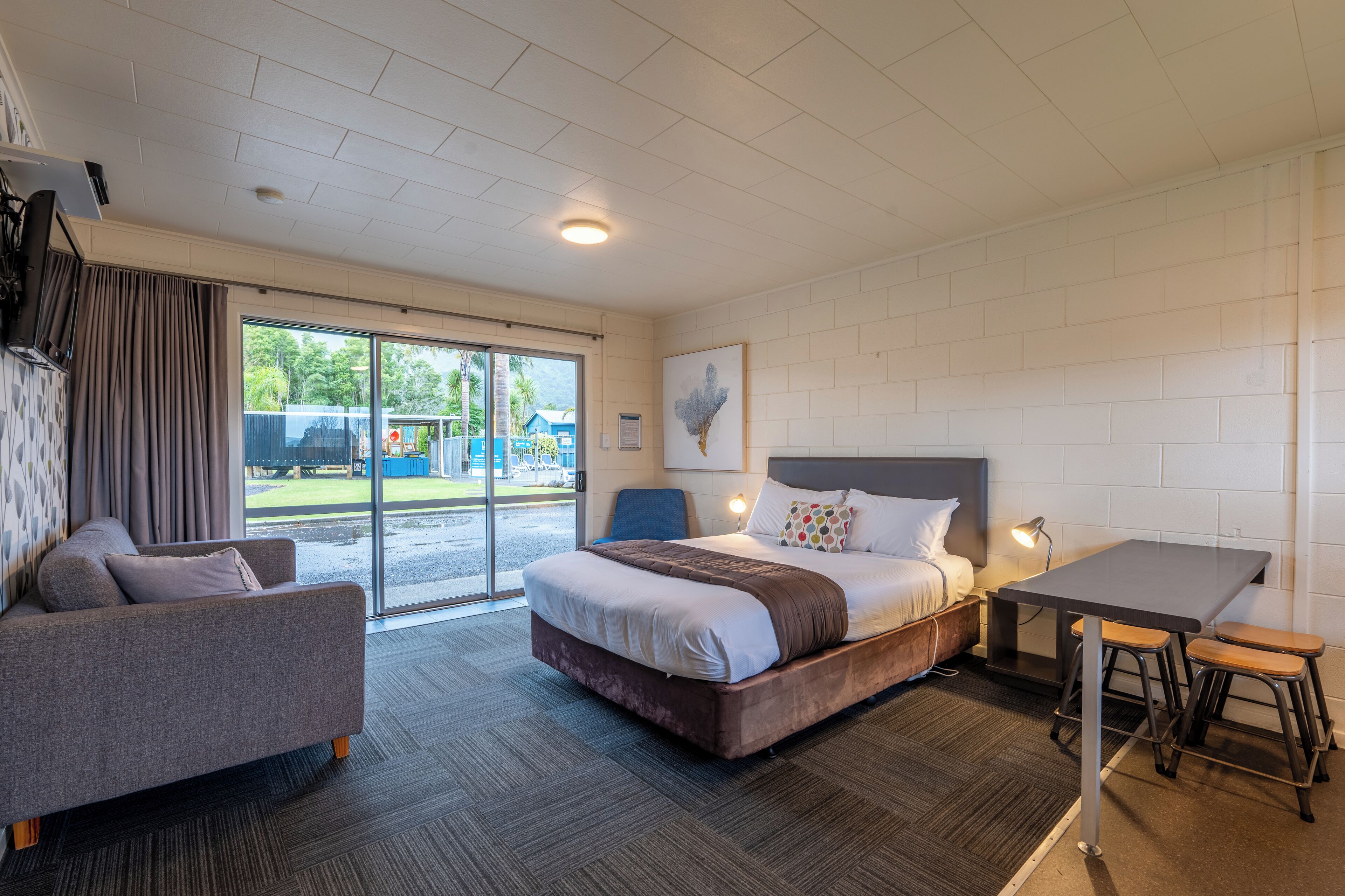 Una habitación de hotel moderna con una cama grande, un sofá, una mesa de comedor con taburetes y vistas al exterior a través de la puerta corredera de vidrio.