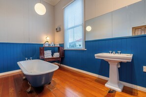 A bathroom with a clawfoot bathtub, a pedestal sink, wooden shelves with towels, and a window with blinds.