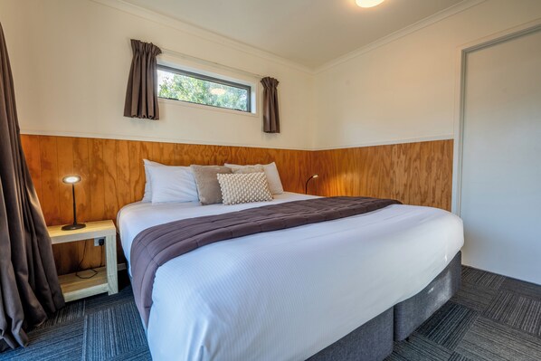 A neatly made bed with a wooden headboard, a bedside table with a lamp, and a window with curtains. - Tasman Holiday Parks – Coromandel (Coromandel)