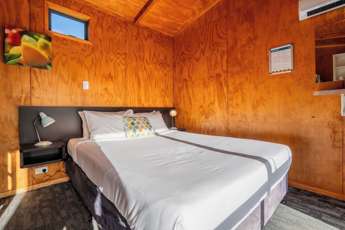 A wooden-paneled room with a bed, a nightstand, a desk, and a small window.