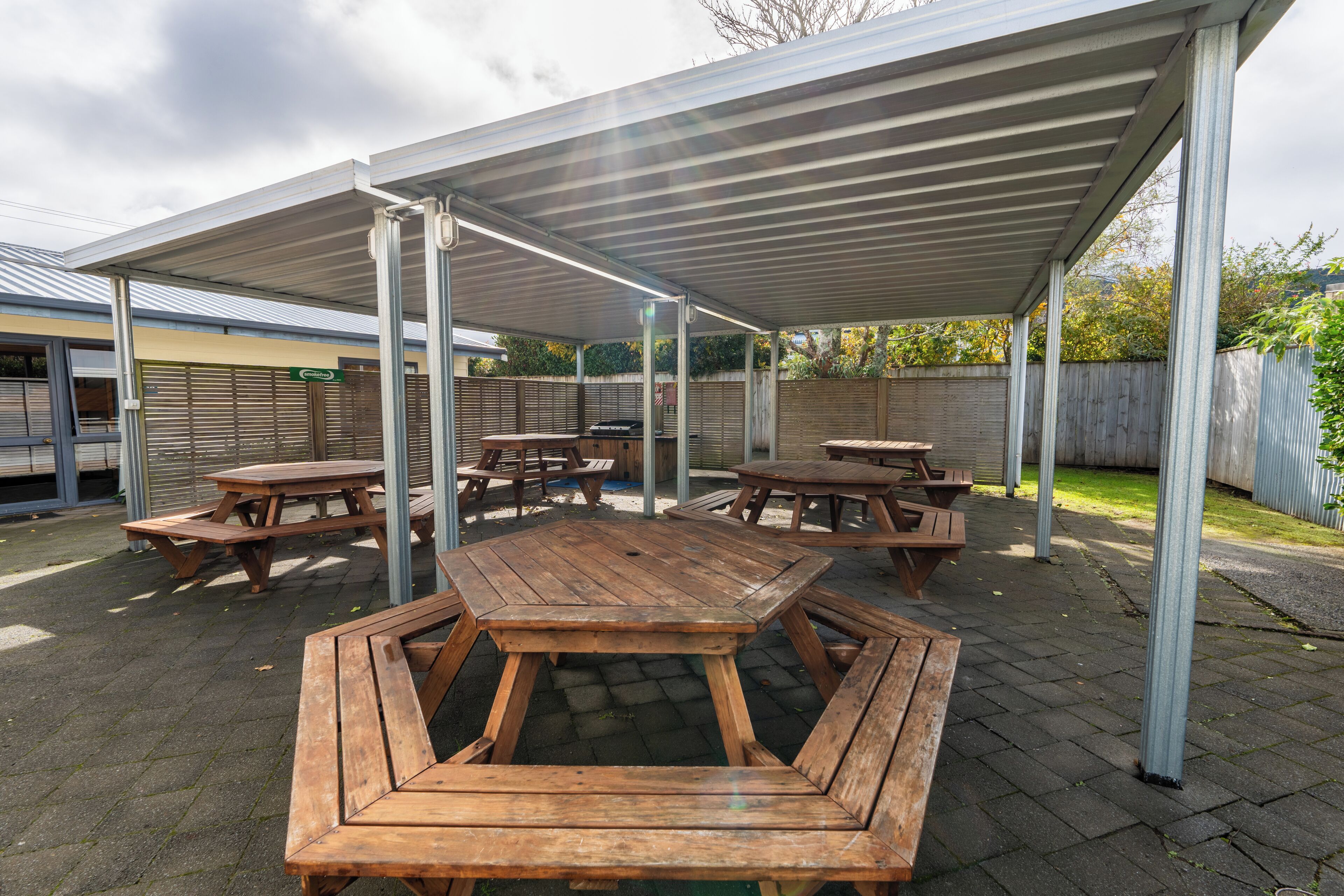 An outdoor patio area with multiple wooden picnic tables and benches, covered by a metal roof.