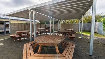 An outdoor patio area with multiple wooden picnic tables and benches, covered by a metal roof.