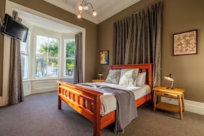 A bedroom with a wooden bed, a nightstand with a lamp, a TV mounted on the wall, and a window with curtains.