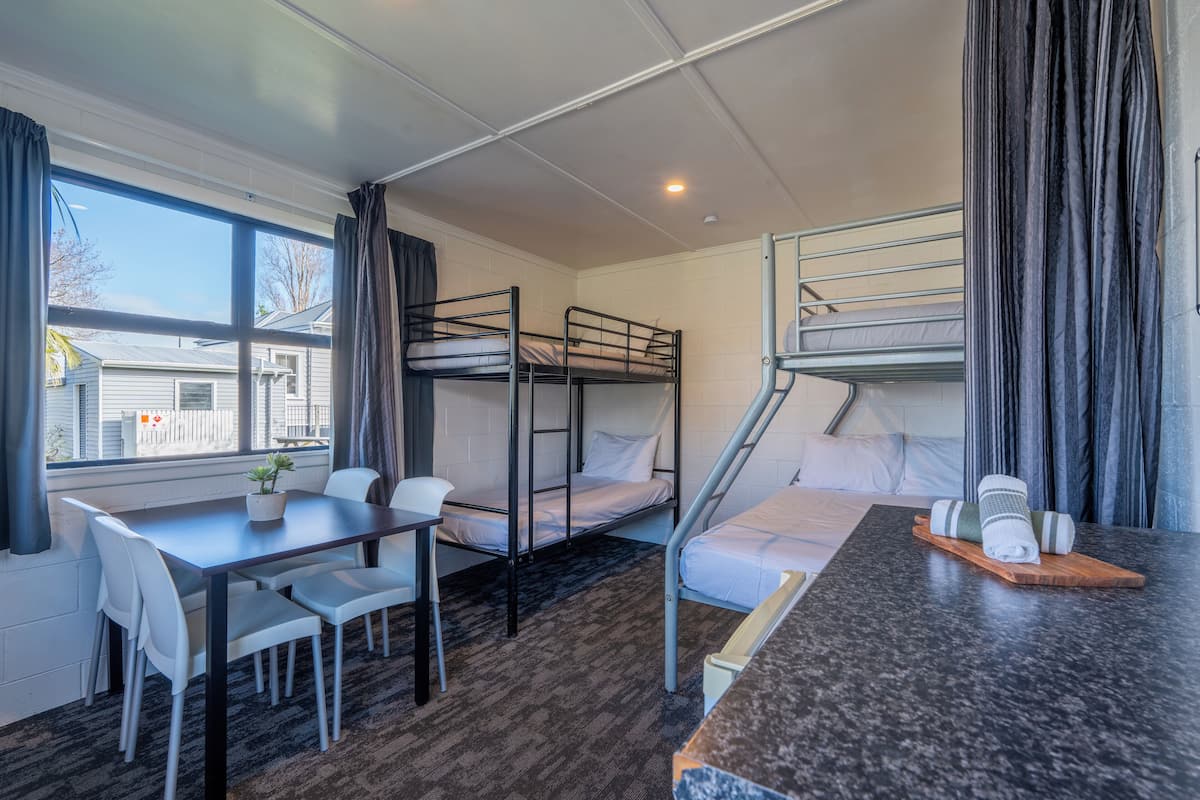 A dormitory room with bunk beds, a desk, chairs, a small table with a plant, and a window with curtains.