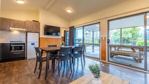 A modern kitchen with wooden cabinets, a built-in microwave, and a refrigerator. There is a dining area with a wooden table and chairs, and a large window offering a view of the outdoors. - Tasman Holiday Parks – Coromandel (Coromandel)