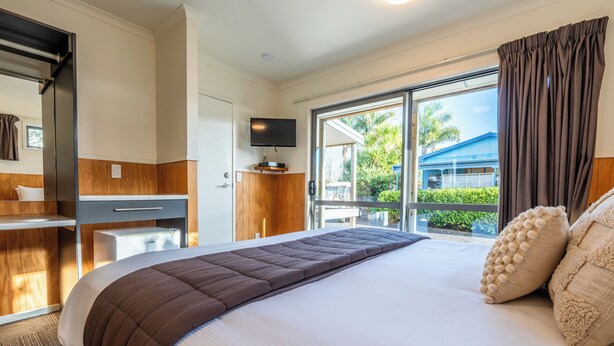 A hotel room with a large bed, a flat-screen TV mounted above it, a desk with a chair, and a window with a view of greenery and a building outside. - Tasman Holiday Parks – Coromandel (Coromandel)